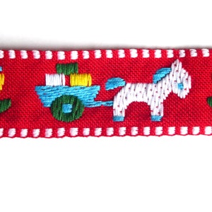 May include: Red fabric ribbon with a white horse pulling a blue wagon with yellow and green blocks, and yellow flowers with green stems on either side.