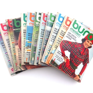 Könnte beinhalten: Ein Stapel alter Burda Moden Magazine mit bunten Covern, die Modeillustrationen und Text in deutscher Sprache zeigen. Das oberste Magazin hat ein rot-grünes Karomuster und den Text "Die Erste Frühlings-Mode" auf dem Cover.