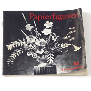 Puede incluir: Una fotograf&iacute;a en blanco y negro de un libro titulado "Papierfiguren" con el n&uacute;mero 52 y el texto "Brummer-Reihe" en la portada. El libro presenta una fotograf&iacute;a en blanco y negro de un jarr&oacute;n con flores.