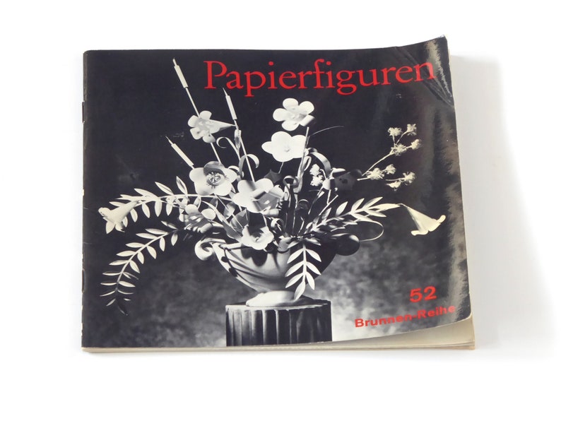 Puede incluir: Una fotograf&iacute;a en blanco y negro de un libro titulado "Papierfiguren" con el n&uacute;mero 52 y el texto "Brummer-Reihe" en la portada. El libro presenta una fotograf&iacute;a en blanco y negro de un jarr&oacute;n con flores.