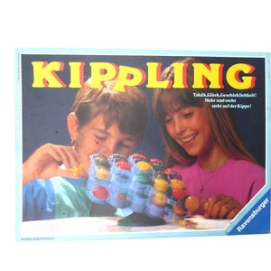Könnte beinhalten: Ein Brettspiel namens "Kippling" mit einem blauen und weißen Spielbrett und bunten Spielsteinen. Die Spielschachtel zeigt zwei Kinder, die das Spiel spielen. Der Text auf der Schachtel lautet "Taktik, Glück, Geschicklichkeit! Mehr und mehr steht auf der Kippe!"