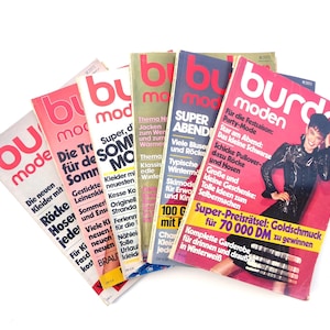 Può includere: Una pila di riviste vintage Burda Moden, con copertine colorate con testo e immagini. Le riviste mostrano segni di usura, con titoli e modelle di copertina visibili. Le riviste sono a ventaglio su una superficie bianca.