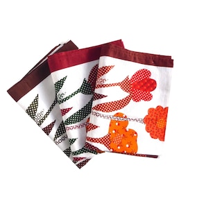 Puede incluir: Tres toallas de cocina blancas con un patrón floral. Las flores son rojas, naranjas y verdes. Las toallas tienen un borde marrón.