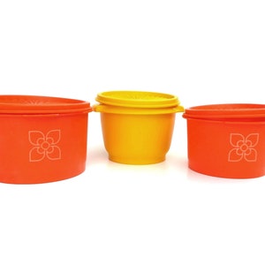 Può includere: Tre contenitori di stoccaggio in plastica con coperchi. I contenitori sono arancioni e gialli con un motivo floreale bianco.