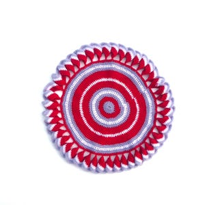 May include: A round, crocheted item with a spiral design. The center is red, with white and gray concentric circles. The outer edge features a scalloped border in red and gray. The item is set against a white background.