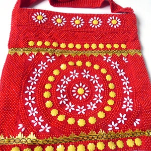 K&ouml;nnte beinhalten: Rote Perlenhandtasche mit einem wei&szlig;en und gelben Blumendesign. Die Tasche hat einen langen Riemen und einen Fransenbesatz.