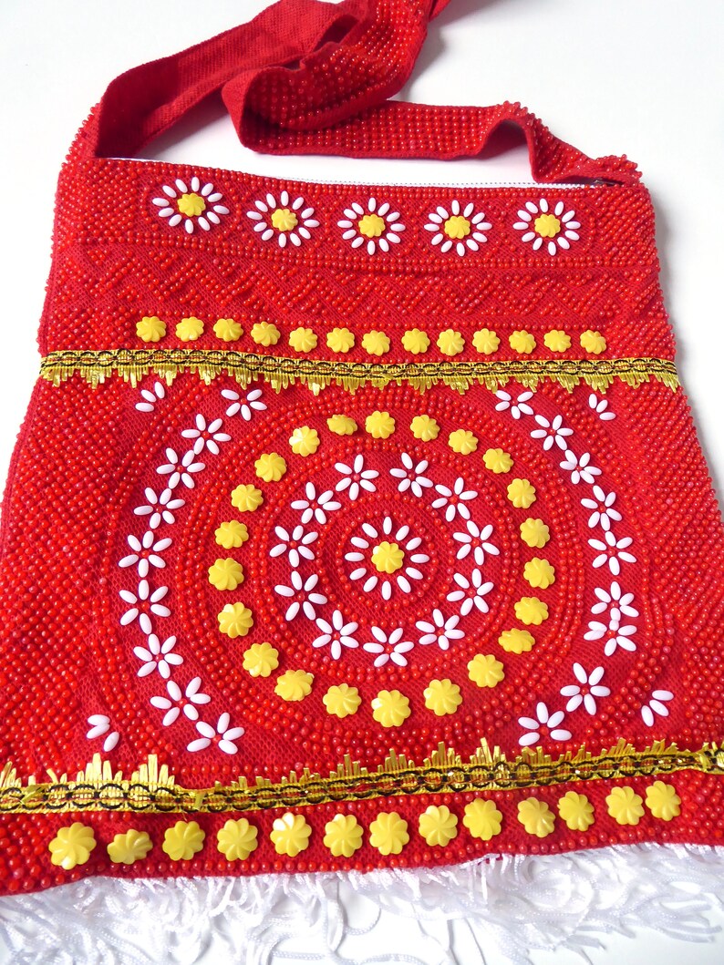 K&ouml;nnte beinhalten: Rote Perlenhandtasche mit einem wei&szlig;en und gelben Blumendesign. Die Tasche hat einen langen Riemen und einen Fransenbesatz.