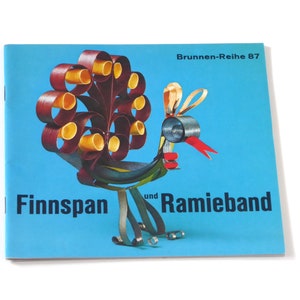 Puede incluir: Una cubierta de libro azul con un dise&ntilde;o de pavo real colorido hecho de papel. El t&iacute;tulo del libro es "Finnspan und Ramieband" y el subt&iacute;tulo es "Brunnen-Reihe 87".