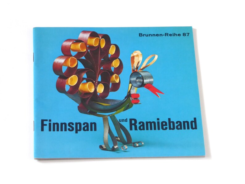 Puede incluir: Una cubierta de libro azul con un dise&ntilde;o de pavo real colorido hecho de papel. El t&iacute;tulo del libro es "Finnspan und Ramieband" y el subt&iacute;tulo es "Brunnen-Reihe 87".