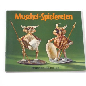 Puede incluir: Un libro titulado "Muschel-Spielereien" con una cubierta verde e ilustraciones de dos figuras hechas de conchas. El libro forma parte de la serie "Brunnen-Reihe 122".