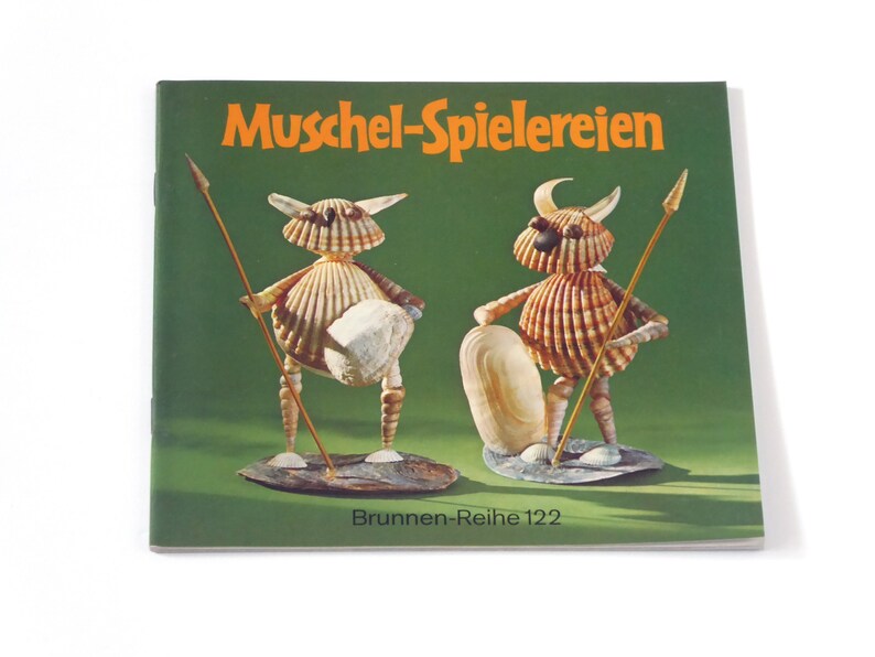 Puede incluir: Un libro titulado "Muschel-Spielereien" con una cubierta verde e ilustraciones de dos figuras hechas de conchas. El libro forma parte de la serie "Brunnen-Reihe 122".