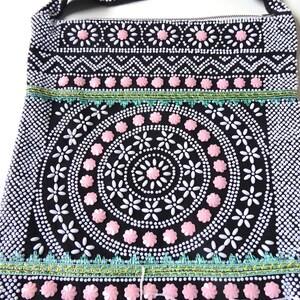 K&ouml;nnte beinhalten: Eine schwarze Perlenhandtasche mit einem rosa-wei&szlig;en Blumendesign. Die Tasche hat einen langen Riemen und einen Fransenbesatz.