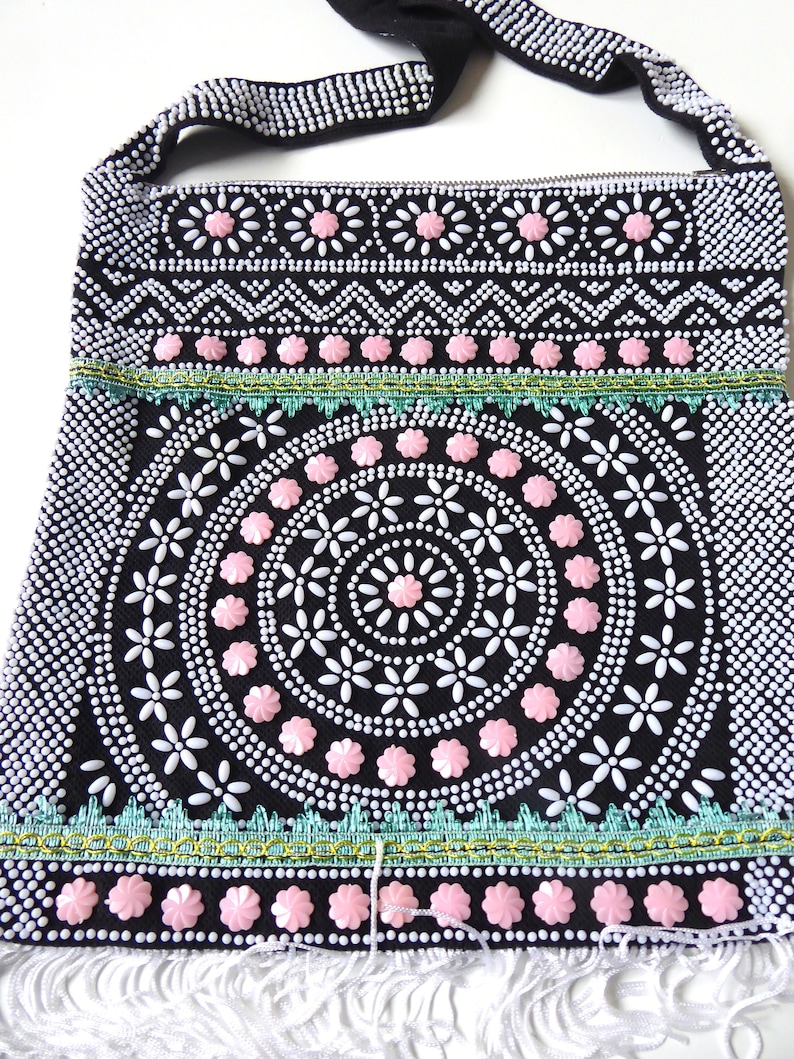 K&ouml;nnte beinhalten: Eine schwarze Perlenhandtasche mit einem rosa-wei&szlig;en Blumendesign. Die Tasche hat einen langen Riemen und einen Fransenbesatz.