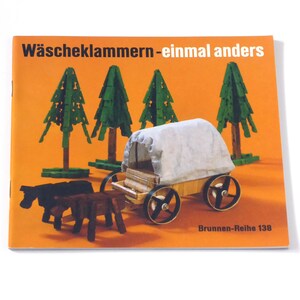 Puede incluir: Una portada de libro con el t&iacute;tulo "W&auml;scheklammern-einmal anders" en texto negro sobre fondo naranja. La portada presenta un carro de madera en miniatura tirado por dos caballos, con una lona blanca. Hay cuatro &aacute;rboles verdes en el fondo. El texto "Brunnen-Reihe 138" est&aacute; en la parte inferior de la portada.