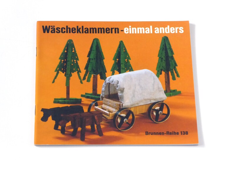 Puede incluir: Una portada de libro con el t&iacute;tulo "W&auml;scheklammern-einmal anders" en texto negro sobre fondo naranja. La portada presenta un carro de madera en miniatura tirado por dos caballos, con una lona blanca. Hay cuatro &aacute;rboles verdes en el fondo. El texto "Brunnen-Reihe 138" est&aacute; en la parte inferior de la portada.