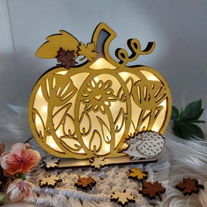 Könnte beinhalten: Eine holzgeschnitzte Kürbis-Lampe mit filigranen Ausschnitten, beleuchtet von warmweißen Lichtern. Mit einem Igel und Blättern. Perfekt für die Herbstdekoration.