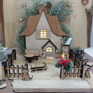 May include: A wooden Christmas village scene with a house, dog figures, a bench, and a sled with the text "Let it Snow." The house has a lighted window and a mailbox with the word "Post."