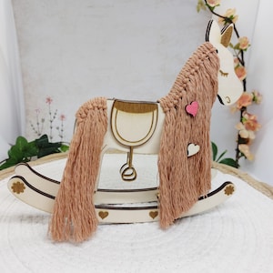 Könnte beinhalten: Ein Holzschaukelpferd mit hellbrauner Mähne und Schweif und einem cremefarbenen Körper. Das Pferd hat einen Sattel und ist mit kleinen Herzen verziert. Mähne und Schweif bestehen aus einem weichen, strukturierten Material. Das Pferd steht auf einer weißen Oberfläche.