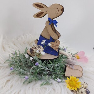 Op de afbeelding: Houten paashaasfiguur met blauwe accenten, een blauwe strik en een hartvormig label. Het konijn is omgeven door groen, bloemen en gespikkelde eieren. Het label luidt "Ich hab DICH lieb".