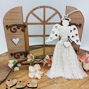 Könnte beinhalten: Ein Holzengel-Ornament mit herzförmigen Ausschnitten auf den Flügeln und einem Heiligenschein. Der Engel hat ein weißes Quastenkleid und einen Herzanhänger. Der Hintergrund zeigt ein Holzfenster und dekorative florale Akzente.