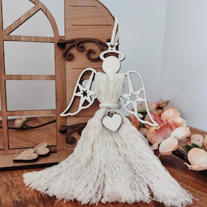 以下が含まれることがあります： 木製の天使のオーナメント。ハローと星型の羽が付いています。天使の体はクリーム色のマクラメでできており、小さな木製のハートが腰からぶら下がっています。オーナメントは、窓のデザインが施された木製の背景に配置されています。