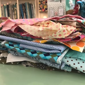 Könnte beinhalten: Ein Stapel bunter Stoffreste mit verschiedenen Mustern wie Punkten, Erdbeeren und floralen Designs. Die Stoffe umfassen Farbtöne von Rosa, Blau, Grün und Braun, was auf ein Bastel- oder Quiltprojekt hindeutet. Die Stoffe sind auf einer hellgrünen Oberfläche gestapelt.