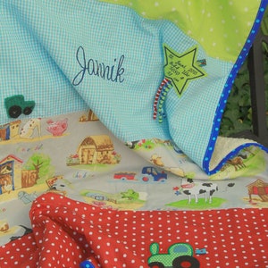Op de afbeelding: Een rood, wit en blauw babydekentje met een groen tractorontwerp. Het dekentje heeft een blauw-wit geruit patroon, een groen-wit sterrenpatroon en een rood-wit stippenpatroon. Het dekentje heeft ook een stervormig label met de tekst "12. Juni 2015 8:44 Uhr 3050 51 cm" en de naam "Jamik" geborduurd erop.