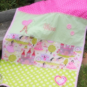 Op de afbeelding: Een roze en groene babydeken met een prinsessen thema. De deken heeft een groen ruitjespatroon met witte stippen, een roze stippenpatroon en een prinsessen-thema stof met een kasteel en een koets. De deken is gepersonaliseerd met de naam "Mina".
