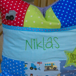 Può includere: Un cuscino patchwork blu e verde con pois bianchi e stelle. Il cuscino ha un pannello centrale a quadri blu e bianco con il nome "Niklas" ricamato in verde. Il cuscino è decorato con un piccolo camion giocattolo grigio e un autobus scolastico giallo.