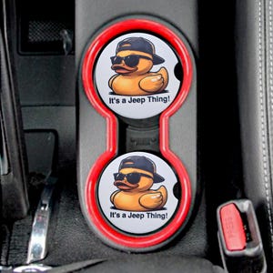 May include: Two red and white car cup holders with a black and yellow rubber ducky design. The ducky is wearing sunglasses and a hat. The text on the cup holder reads "It's a Jeep Thing!"