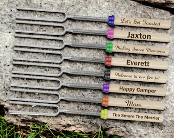 May include: Set of eight personalized marshmallow roasting sticks with wooden handles. The handles are decorated with different colored rings and engraved text, including "Let's Get Toasted", "Jaxton", "Making Smore Memories", "Everett", "Welcome to our fire pit", "Happy Camper", "Mom", and "The Smore The Merrier".