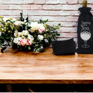 May include: A black wine bottle bag with a white golf ball design and the text "Tasting at the Tee". The bag is sitting on a wooden table with a bouquet of white and pink flowers and a stack of black napkins.
