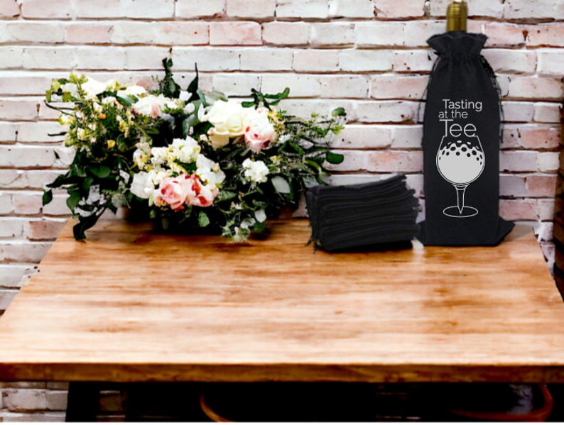 May include: A black wine bottle bag with a white golf ball design and the text "Tasting at the Tee". The bag is sitting on a wooden table with a bouquet of white and pink flowers and a stack of black napkins.