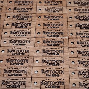 May include: A close-up of multiple wooden tags with the text "Sawtooth Caverns" engraved in black. The tags are arranged in rows and are all identical.