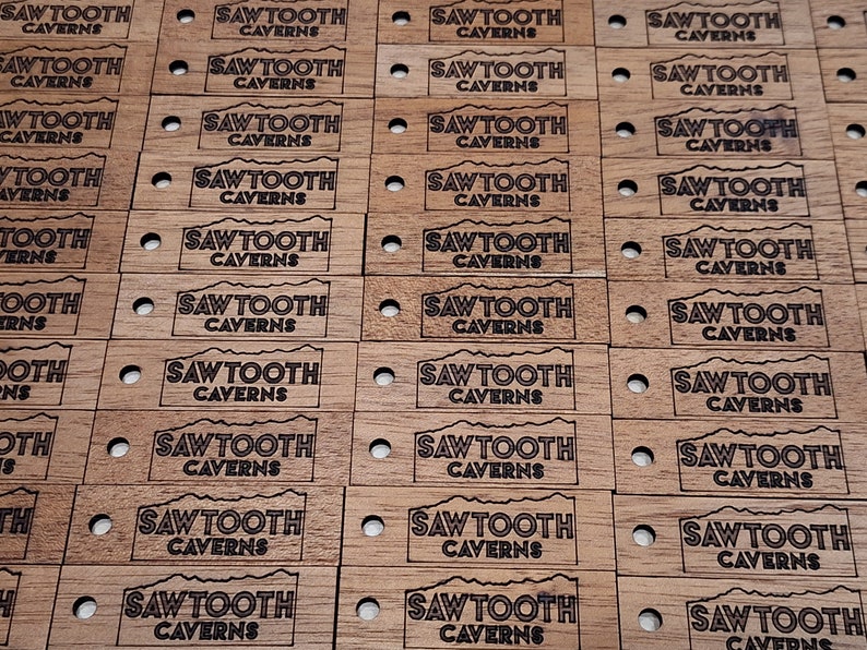 May include: A close-up of multiple wooden tags with the text "Sawtooth Caverns" engraved in black. The tags are arranged in rows and are all identical.