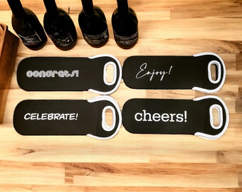 May include: Four black wine bottle carriers with white handles and white text. The text on the carriers reads "Congrats!" "Celebrate!" "Enjoy!" and "Cheers!"