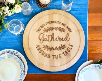 May include: A round wooden serving board with the text "The best memories are made gathered around the table" engraved on it. The board is on a blue tablecloth with white flowers and glasses.