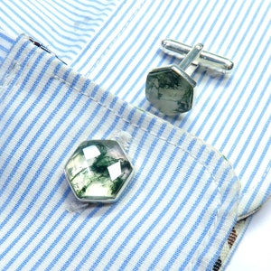Puede incluir: Un par de gemelos de plata con piedras de ágata verde musgo. Los gemelos están sobre una camisa a rayas azules y blancas.