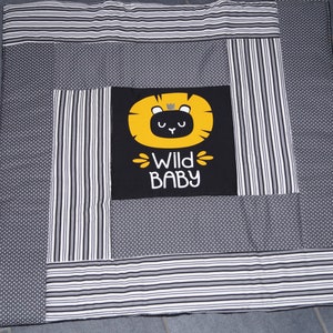 Könnte beinhalten: Eine quadratische Babydecke mit Patchwork-Design. Die Decke hat ein zentrales schwarzes Quadrat mit einer gelben Löwengrafik und den Worten "WILD BABY". Der umgebende Stoff ist grau mit weißen Punkten und Streifen.