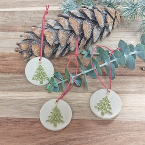 Könnte beinhalten: Drei runde, cremefarbene Keramik-Weihnachtsbaumornamente mit grünen Baumdesigns und roten Aufhängeschnüren. Ein großer Tannenzapfen und Eukalyptuszweige befinden sich im Hintergrund, alles auf einer Holzoberfläche.