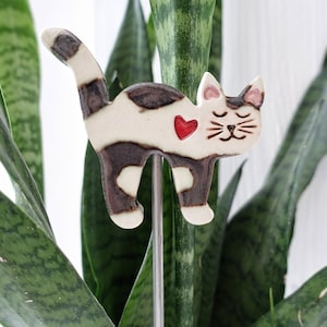 Könnte beinhalten: Ein Pflanzenstecker in Form einer Katze aus Keramik. Die Katze hat einen weißen Körper mit braunen Flecken und einem roten Herz. Sie hat geschlossene Augen und ein lächelndes Gesicht. Der Metallstab steckt in einer grünen Pflanze.