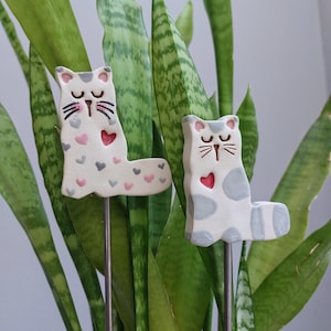 Könnte beinhalten: Zwei Keramik-Katzen-Pflanzstäbe. Die Katzen sind weiß mit grauen und rosa Herz- und Punktmustern. Jede Katze hat ein rotes Herz auf der Brust und einen Metallstab zum Einsetzen in die Erde.