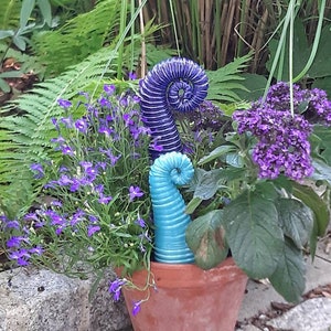 Könnte beinhalten: Ein Terrakotta-Topf gefüllt mit lila Blumen, grünen Farnen und zwei dekorativen Glasskulpturen. Eine Skulptur ist eine hohe, spiralförmige blaue Form, die andere eine kürzere, türkisfarbene Spirale. Die Anordnung steht vor üppigem Grün.