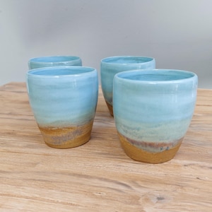 Könnte beinhalten: Ein Set aus vier Keramikbechern mit einem glatten, hellblau glasierten oberen Teil, der in eine strukturierte, erdfarbene braune Basis übergeht. Jeder Becher hat eine abgerundete, henkellose Silhouette mit subtilen Farbvariationen.