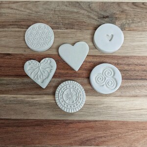 Könnte beinhalten: Sechs weiße Keramik-Ausstecher mit verschiedenen Designs: eine Blume, ein Herz, ein Wirbel, eine Blume, ein Herz und ein geometrisches Muster. Die Ausstecher befinden sich auf einer Holzoberfläche.