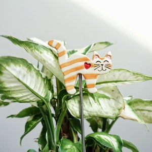 Könnte beinhalten: Eine Keramik-Katzenfigur mit orange-weißen Streifen und einem roten Herzen auf dem Rücken. Die Figur ist an einem Metallstab befestigt und steht in einer grün-weißen Pflanze.