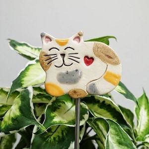 Könnte beinhalten: Eine Keramik-Katzenfigur mit einem lächelnden Gesicht und einem roten Herzen an der Seite. Die Katze ist in Weiß-, Braun-, Gelb- und Grautönen bemalt. Die Figur ist an einem Metallpfahl befestigt, wodurch sie sich als Pflanzenmarke eignet.
