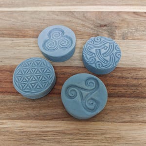 Könnte beinhalten: Vier runde, hellblaue Seifen mit geprägten Designs. Die Designs umfassen eine dreifache Spirale, einen keltischen Knoten, ein Blumen des Lebens-Muster und ein Triskele. Die Seifen sind auf einer Holzoberfläche angeordnet.