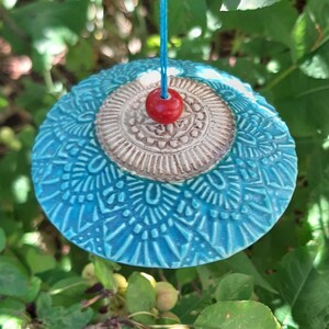 Könnte beinhalten: Handgefertigtes Keramik-Windspiel mit türkisfarbenem, gemustertem Boden und beigefarbenem Zentrum. Eine kleine rote Perle hängt an einer blauen Schnur. Das Windspiel hängt vor einem Hintergrund aus grünen Blättern und kleinen grünen Früchten.