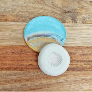 Könnte beinhalten: Ein Keramikuntersetzer mit Strandmotiv, mit blauem Himmel und Sandstrand. Neben dem Untersetzer befindet sich eine weiße, muschelförmige Seifenschale. Die Artikel befinden sich auf einer Holzoberfläche.